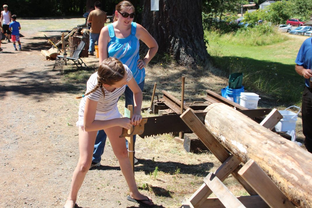 a little girl cuts a large log with a saw while a woman watches her at the Oregon Trails Days in Tenino, WA