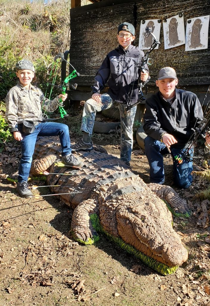 three boys, two with a foot on a fake alligator, pose with their archery bows in hand