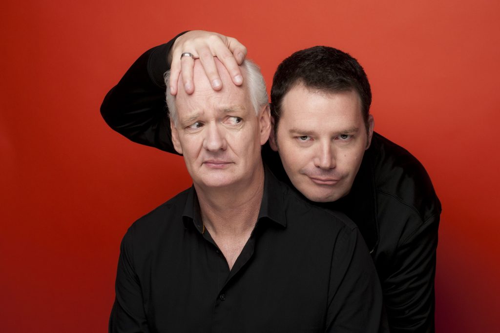 one man with his hand on another man's bald head, while he looks at him sideways against a red background