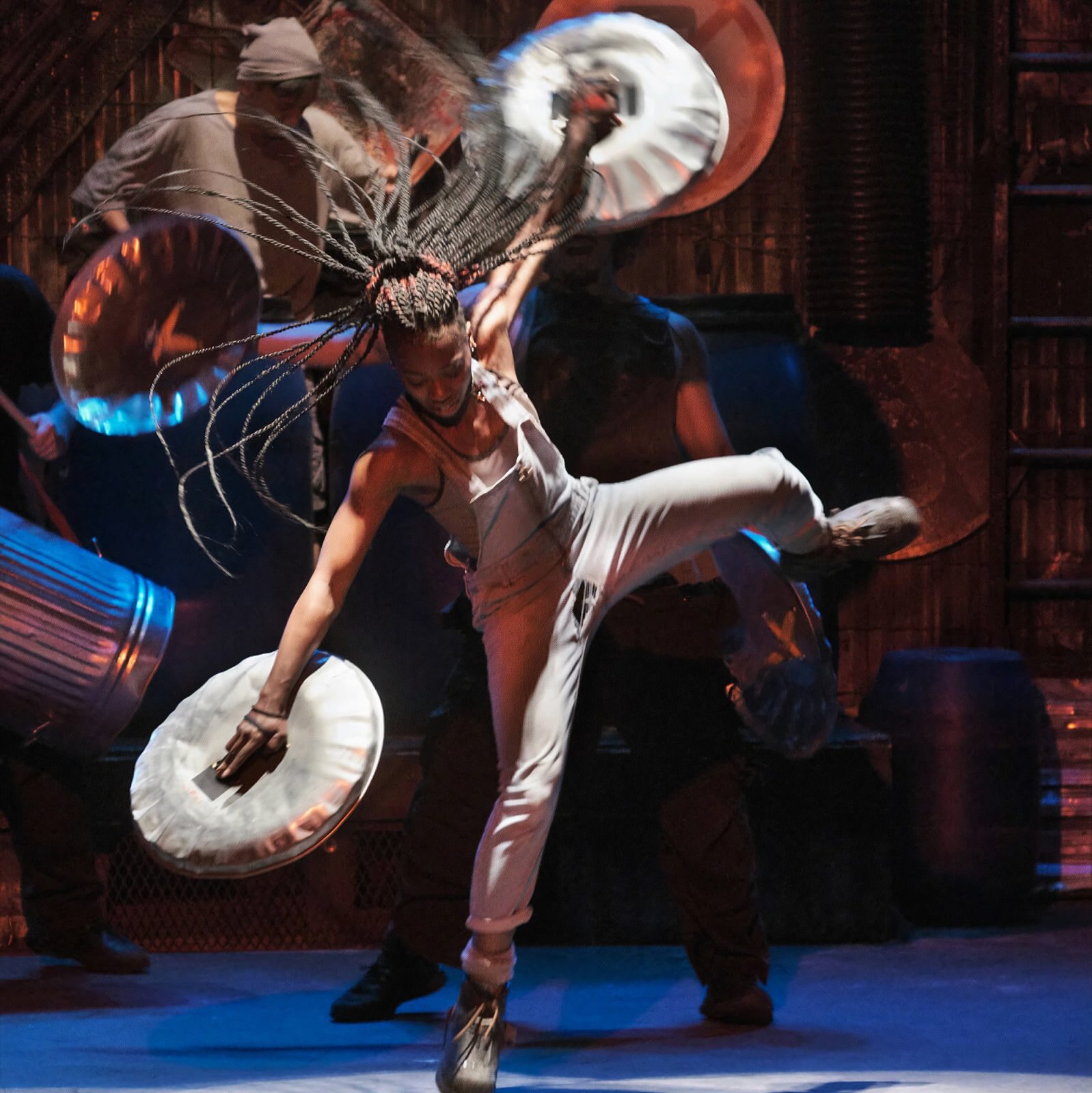 woman with dreaded hair in white overalls with trashcan lids in both sands, jumping around on stage