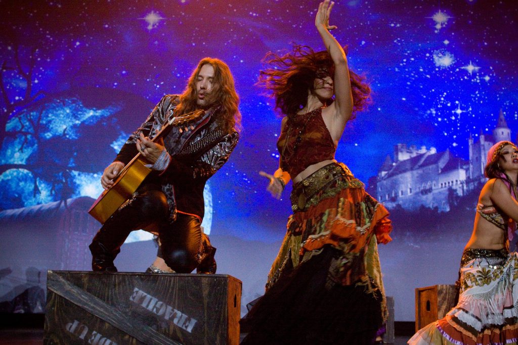 El maestro Benis baila con tonos tierra contra un cielo azul estrellado junto a una mujer que toca la guitarra en el escenario.