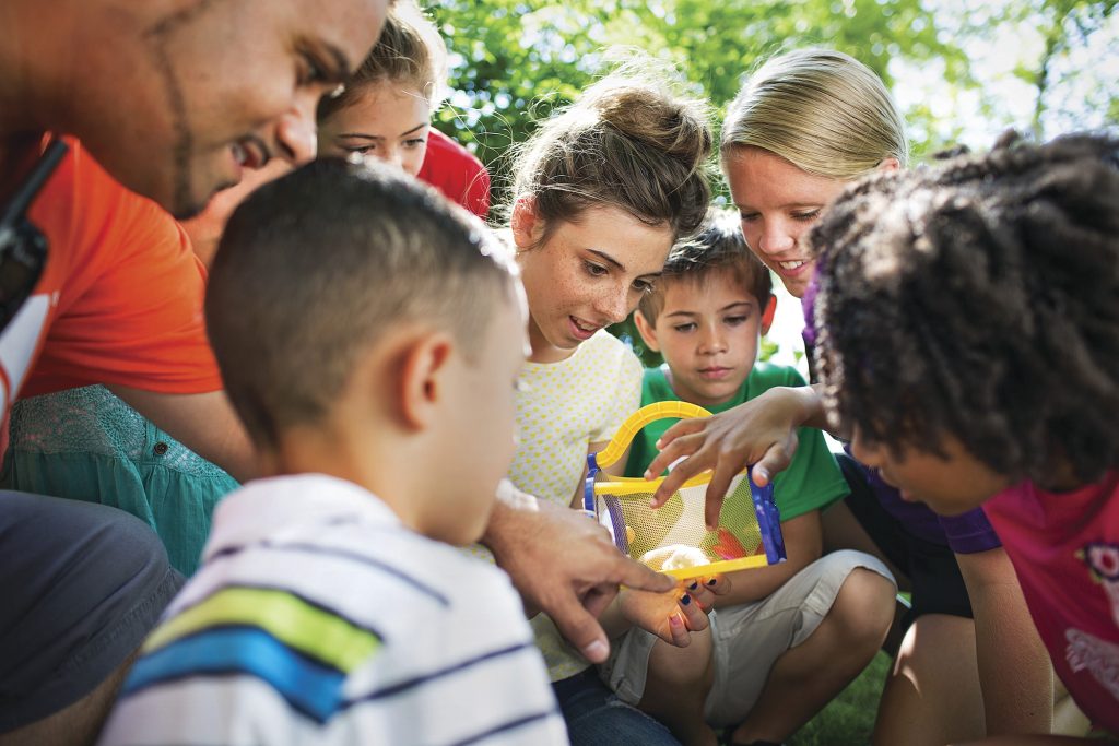 a bunch of kids with a couple adults looking at something in a small yellow and blue mesh cage outside