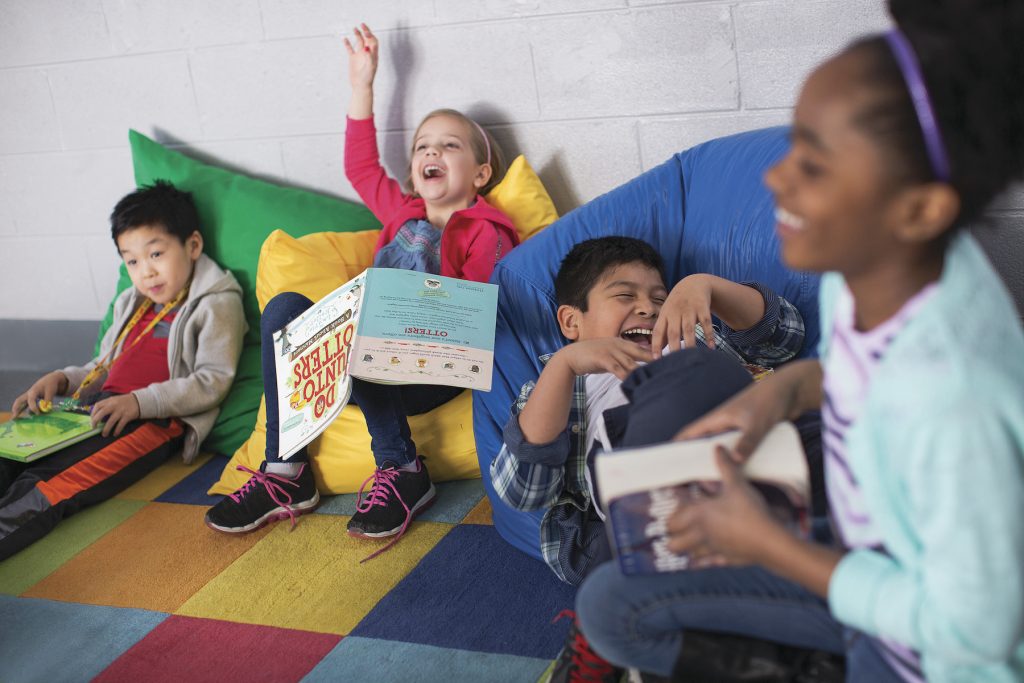 four kids on colorful pillows on a colorful block rug on the floor, reading books and laughing. One girl has her hand raised
