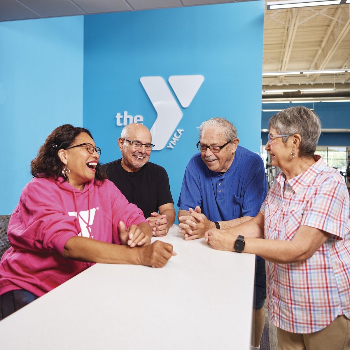 4 senior citizens standing around a white table with a blue wall with the "Y" symbol on it.