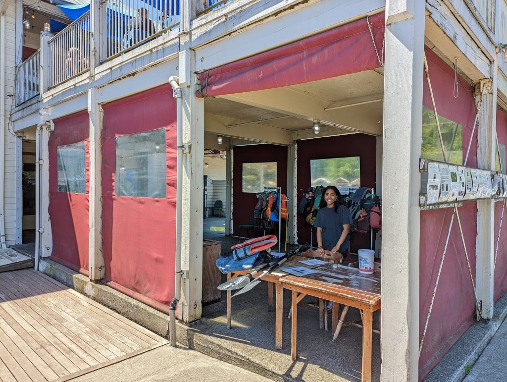 woman at the table at Tugboat Annie's rental kiosk - a red building with a large open walkway with kayaks piled behind the table