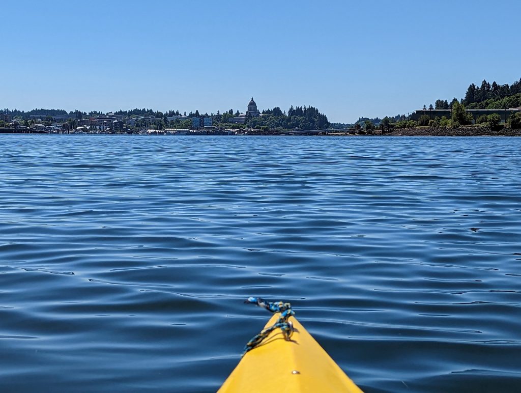 tip of a yellow kayak in the Puget Sound pointed toward the Olympia Capitol, which can be seen from a distance