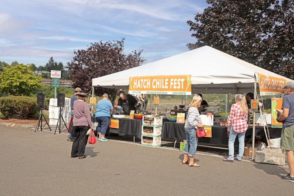 customers mill around a white tent with crates of chilis a banner on the tent says 'Hatch Chile Fest'