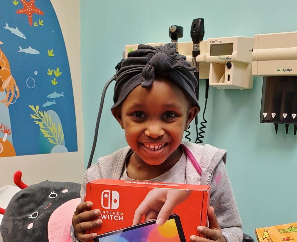 Little girl in a hospital room with a wrap on her head, smiling at the camera holding a Wii Switch
