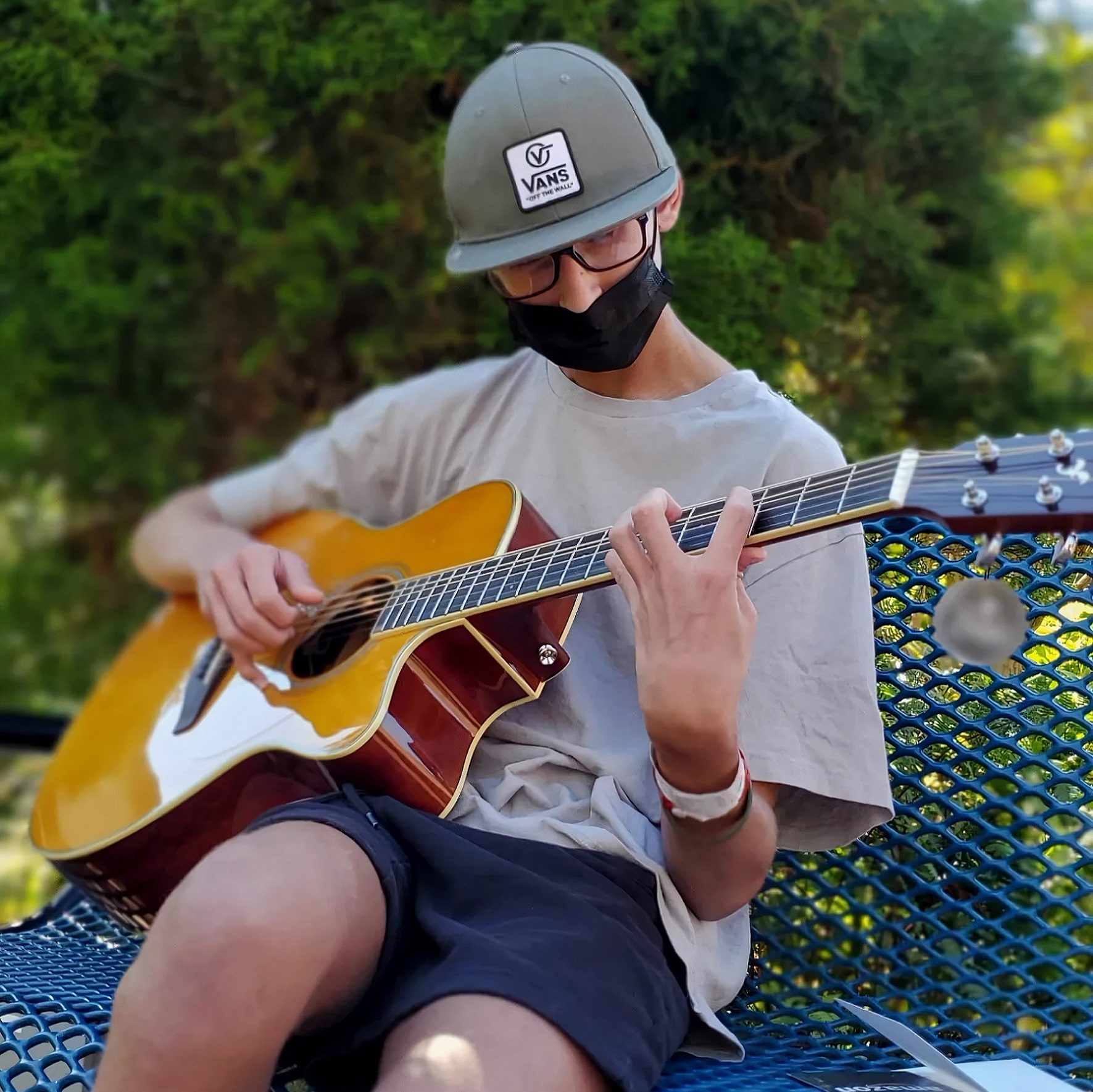 kid sitting on a bench with a cap and a face mask on, playing the guitar. He has a medical bracelet on his wrist.