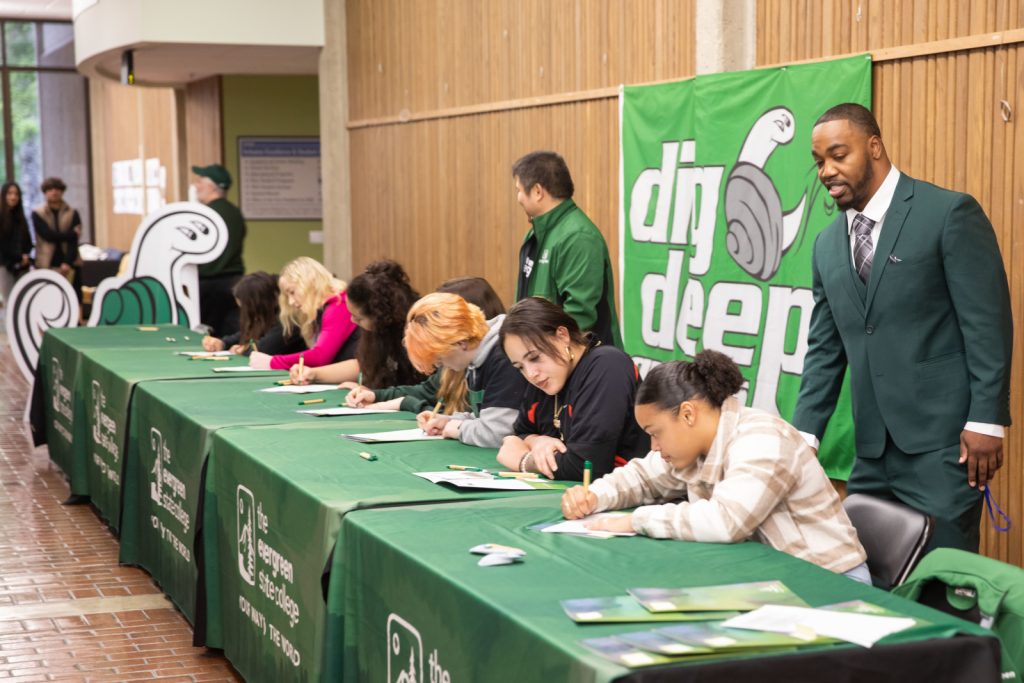 college students signing on to play sports at large tables covered in green cloths, two men stand behind them