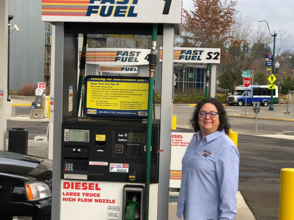 woman with a Fast Fuel shirt on stands by a gas pump, smiling
