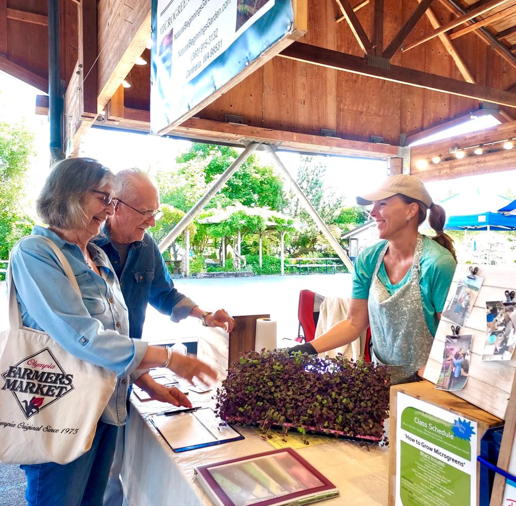 The Fitzgeralds buying microgreens from Kathy Aust at her booth at the Olympia Farmers Market. Kathy is holding a plate of micro greens and smiling across a counter at the Fitzgeralds