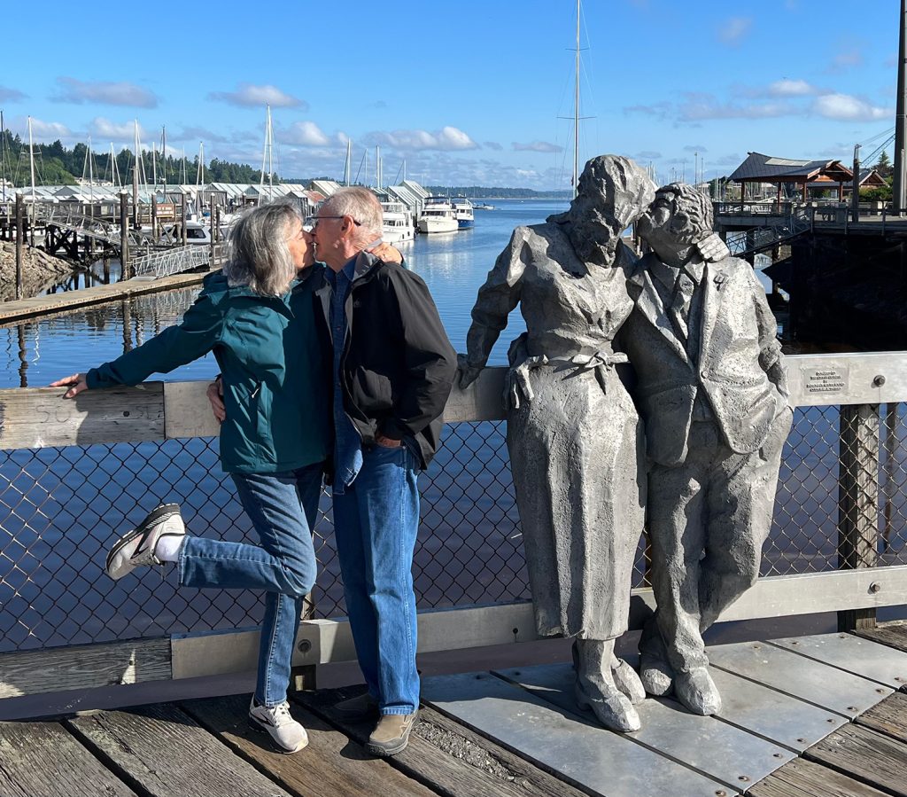 Jeanette and Bill pose for a photo next to a statue of a couple with the Puget Sound behind them.