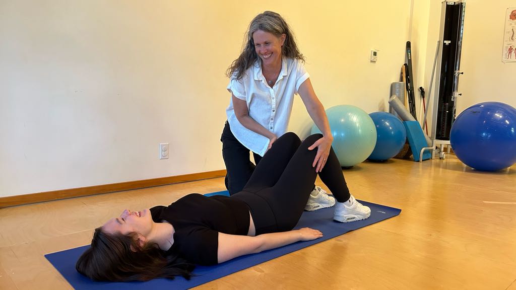 Michelle kneeling next to a patient lying on their back on a mat with their knees bent up. Michelle has her hands on either side of the patient's knees