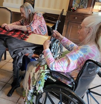 Two women in wheelchairs work on tying blankets on a table