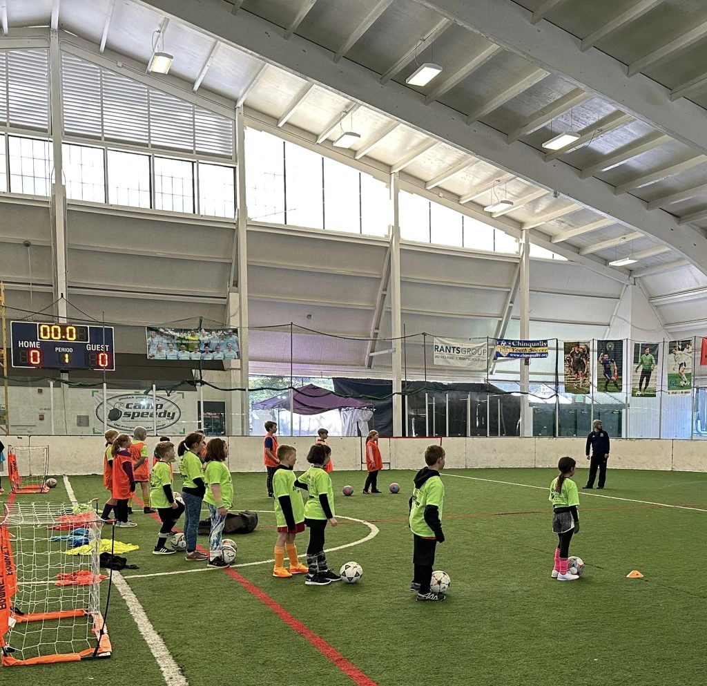 young kids in green an and orange t-shirts stand with soccer balls on an indoor field