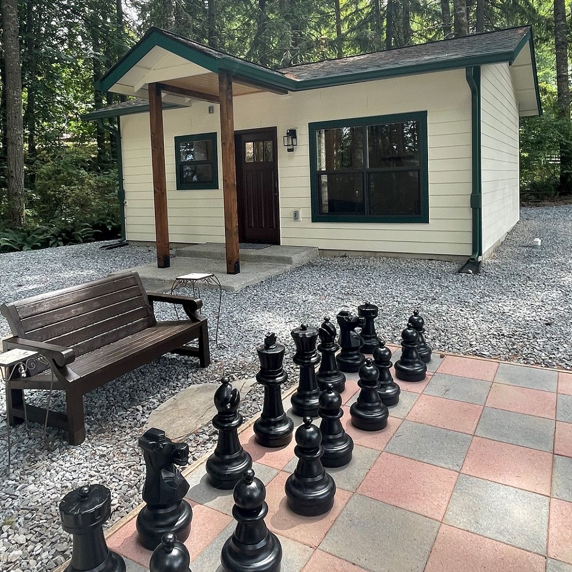 A small additional dwelling unit with a large, oversized chess board and black chess pieces outside. A bench in gravel is next to the board.