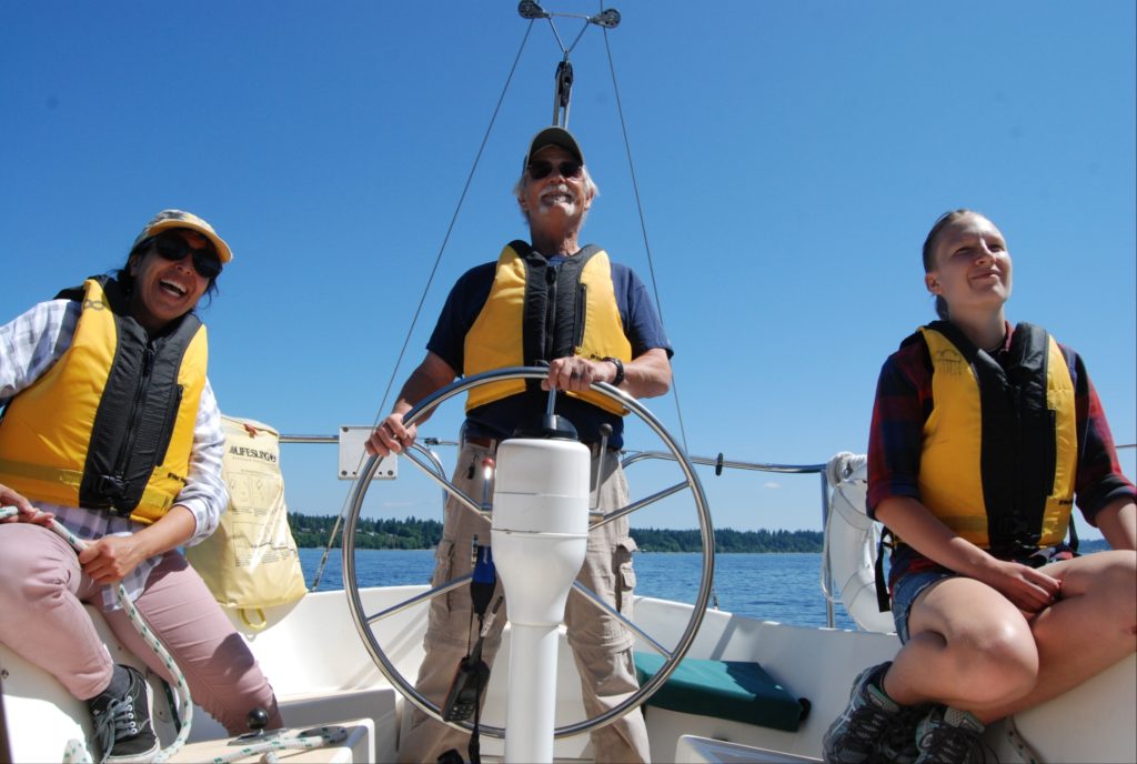 an older man at the wheel of a sail boat, with two women seated on either side of the boat