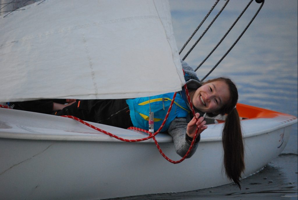 a young girl lying sideway reaching out of a boat holding a red rope attached to a sail