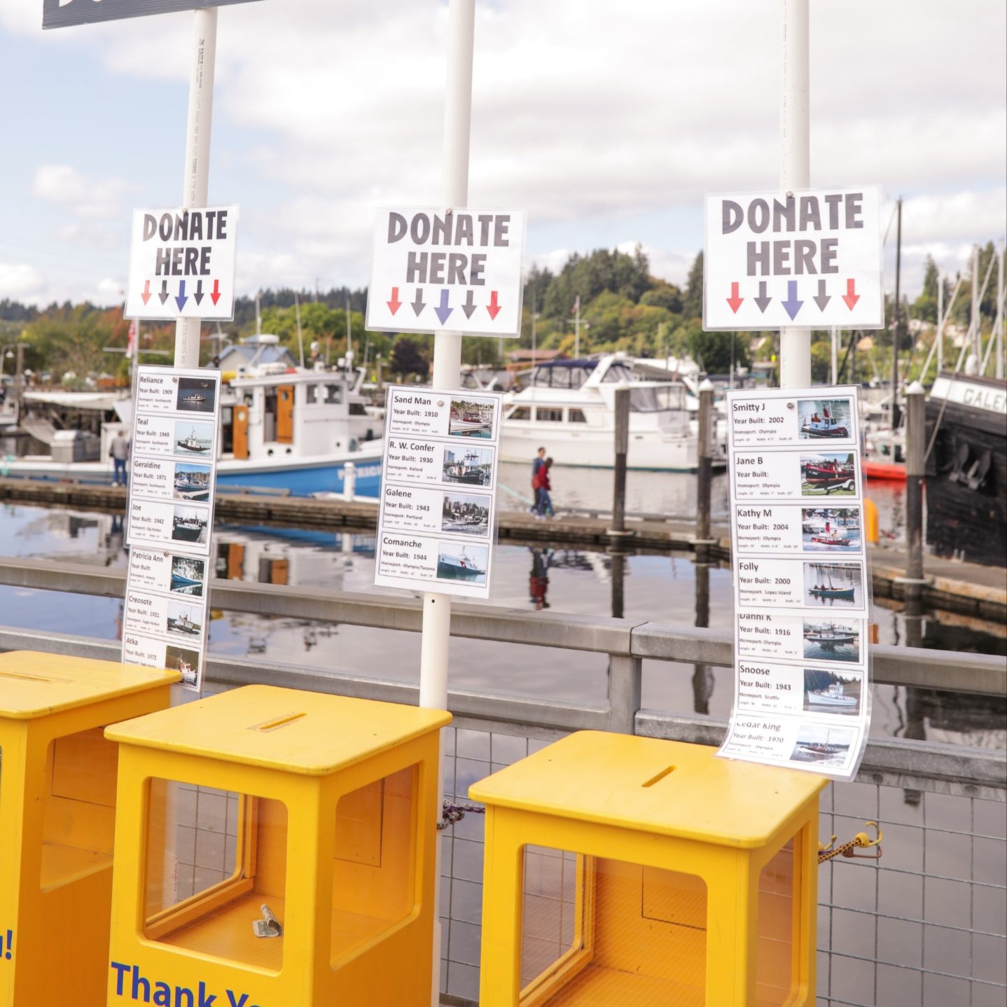 3 Yellow boxes with signs that say, 'donate here' and a large sign above it them that says, 'Donate Here and Thank you!' with the Olympia Harbor Days logo and drawings of tug boats
