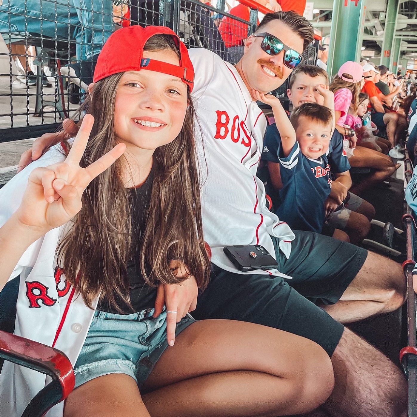 Dr. Hadden and three of this kids in the bleacher at a sports game, posing for the camera. His daughter is making a peace sign