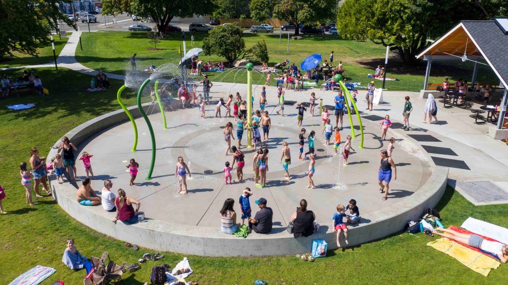 large circular concrete splash pad with tall, green metal spouts sprinkling water. Kids and adults are playing in the water and sitting on the concrete wall around it.