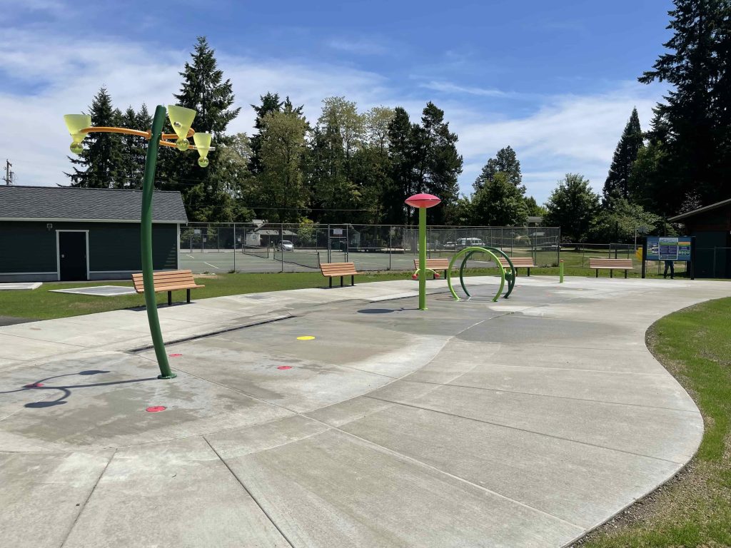 concrete area with benches surrounding it and tennis courts behind it. The concrete splash pad has tall green spouts and one circular one.