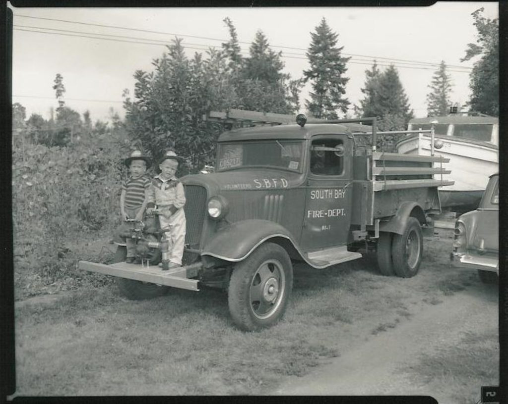 black and white photo of an old South Bay Fire Department truck with two kids standing on it's front bumper