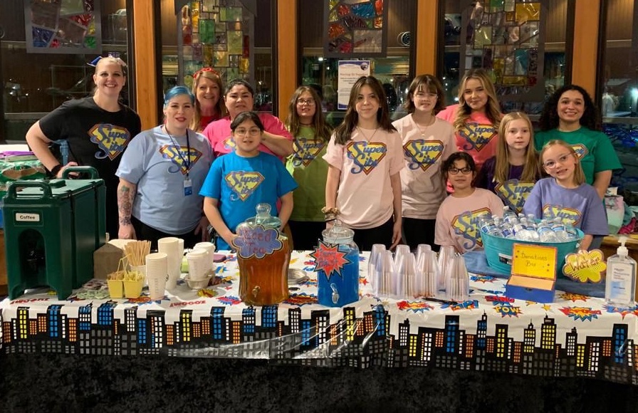 A large group of kids wearing 'super sibs' shirts pose for a photo behind a table with drink coolers, water bottles and plastic cups on it.