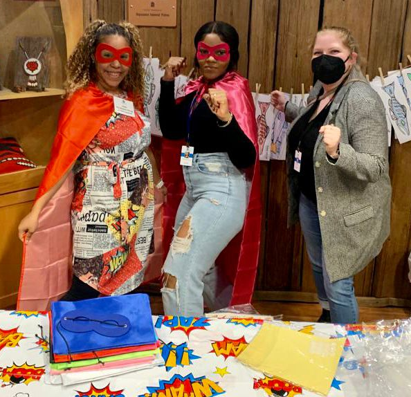 Three women, two dressed up as superheroes, post for a photo behind a table with a fabric on it and a tablecloth that says 'Zap; 'Boom' on it