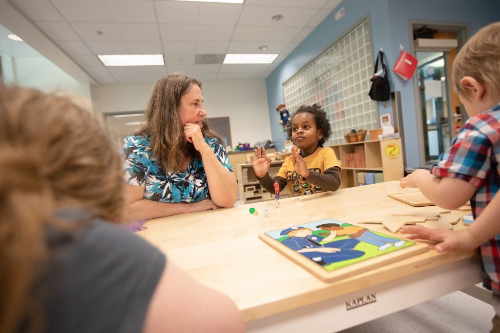 adult sitting at a table with kids in a classroom. There are wooden puzzles and dice on the table