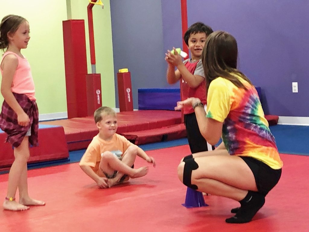 three kids, one sitting on a gym mat, look at an adult that is crouching down.