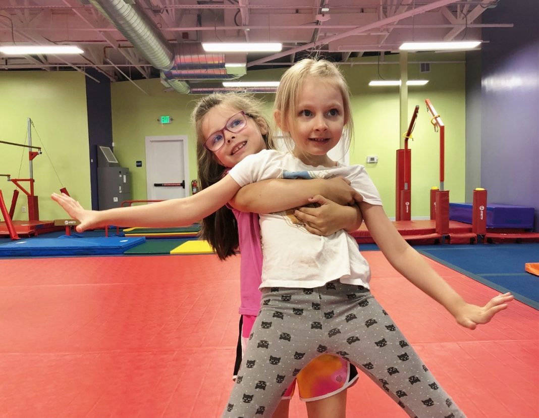 a little girl guys and lifts up another little girl from behind, inside a gym with colored mats in stacks behind them.