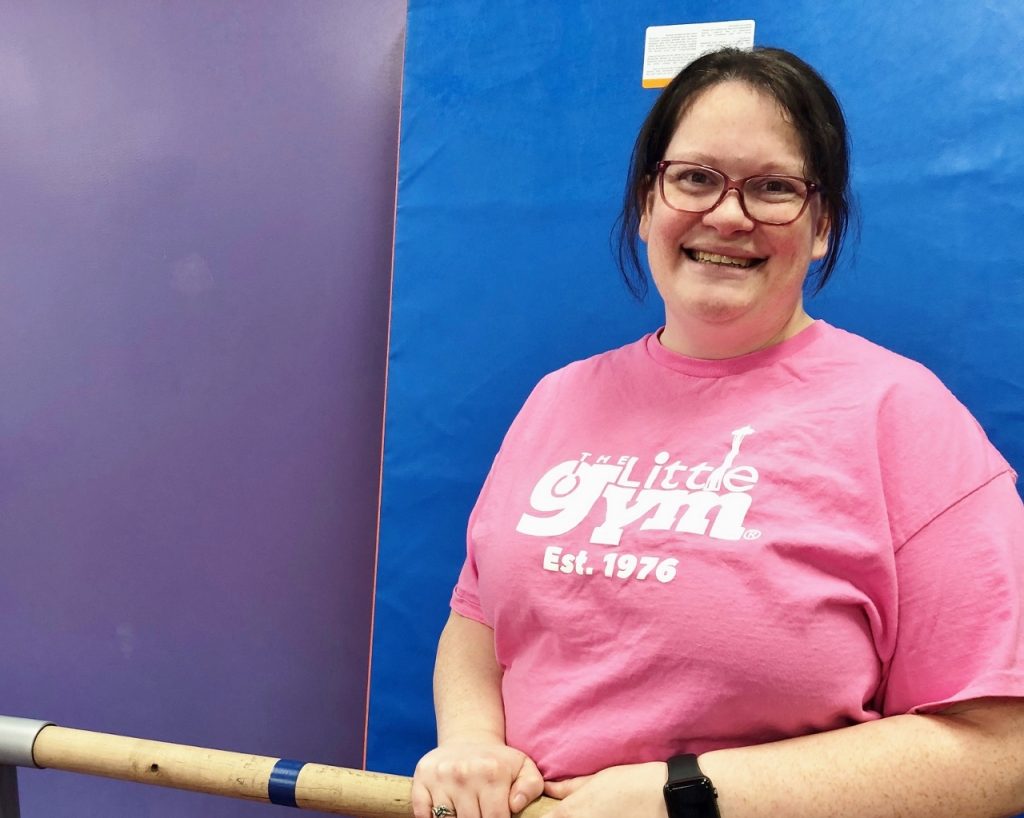Melissa Lilley in a pink shirt that say, 'The Little Gym, est. 1976' smiles for the camera.