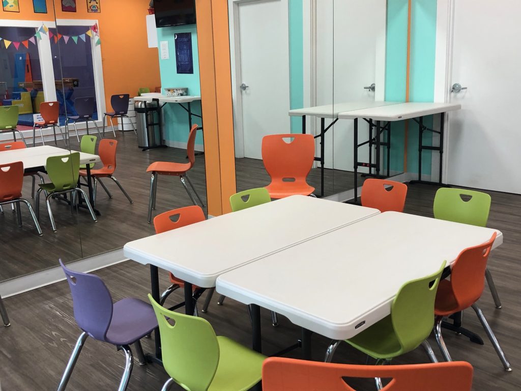 brightly colored room with two white tables pushed together surrounded by orange, green and purple chairs