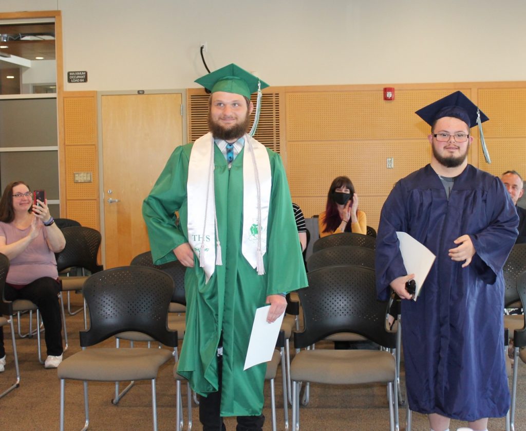 Two men, one in a green graduation cap and gown, one in a blue cap and gown stand in front of chairs, a couple people are sitting in the chair behind them.