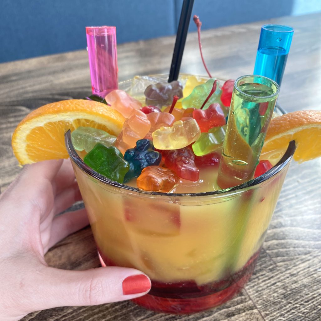 a fancy cocktail in a short glass filled with test tubes of pink, green and yellow, a bunch of gummy bears and two orange slices