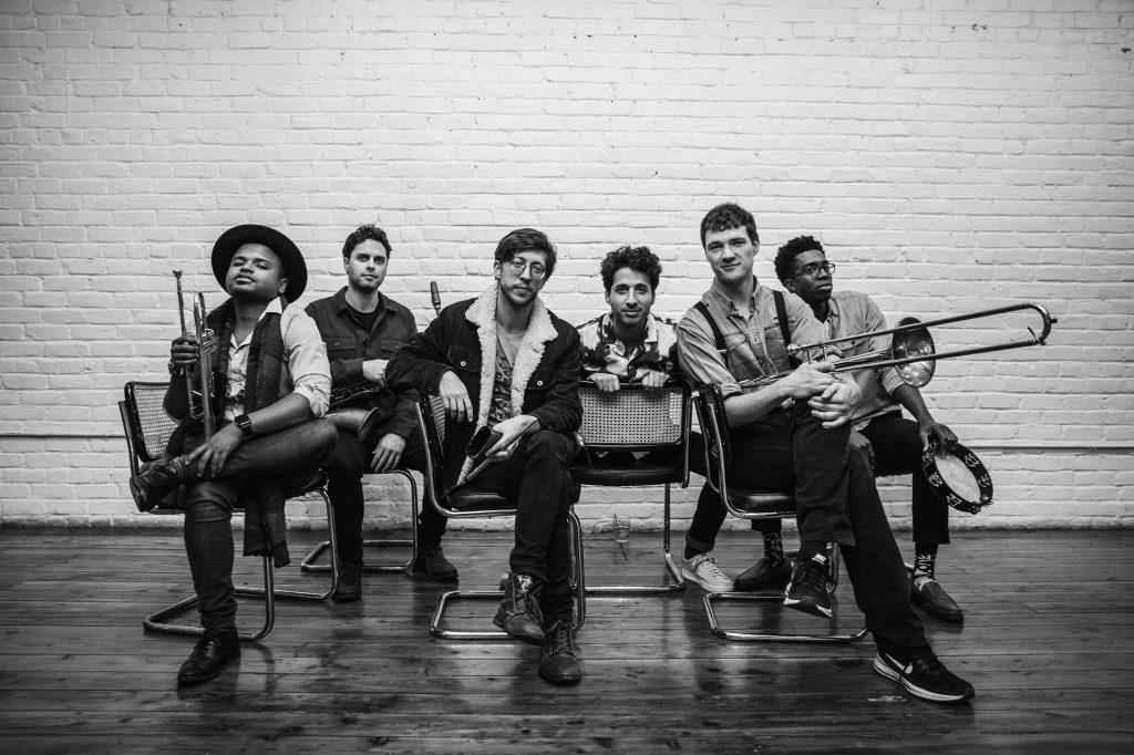 black and white photo of Sammy Miller and the Congregation sitting with their instruments on a bench with a white brick background