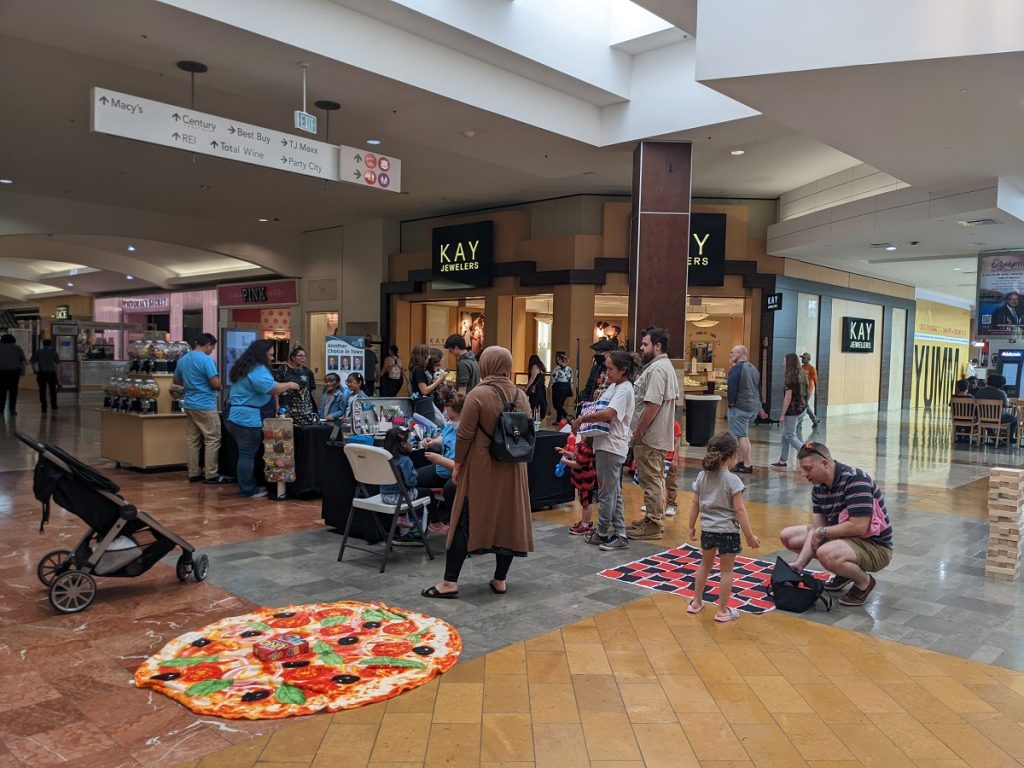 a group of people and kids inside the Capital Mall, some doing face painting, some with balloon twists. A giant pizza made of fabric is on the ground as part of a game