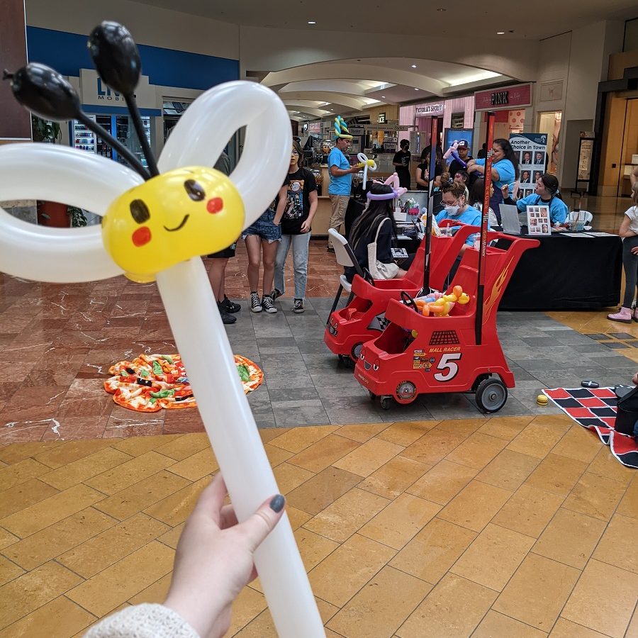 a women holding a balloon twist that looks like a bug on a wand. In the background, families are doing various activities inside the Capital Mall