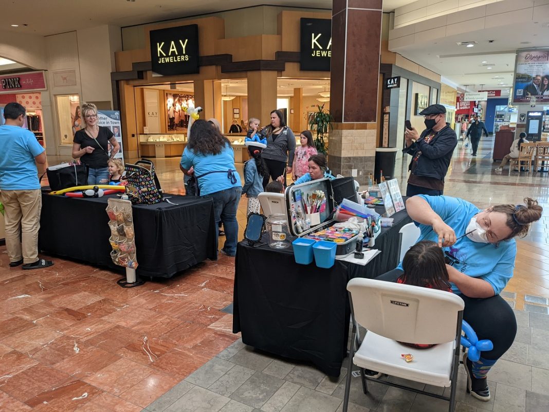 a group of people and kids inside the Capital Mall, some doing face painting, some with balloon twists, at various tables covered with black cloths