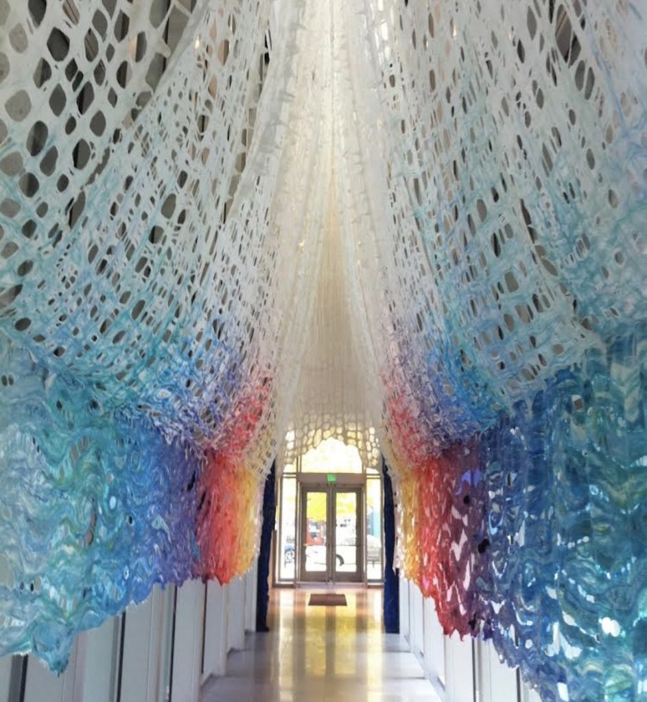 Large felted curtain hanging down from the ceiling on either side of a hallway, starts out white then fades to many colors.