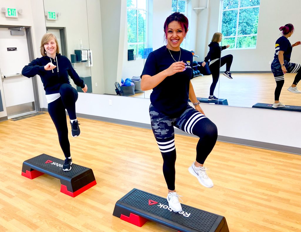 Two women in front of large mirror working out on step ups. 