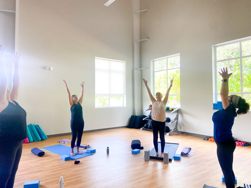 Women stand on yoga mats, arms lifted up. yoga blocks and water bottles are on the floor