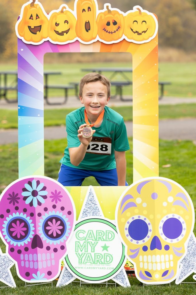 A kid posing with a medal in a cardboard cut out frame with sugar skulls on it that says, 'Card my Yard'