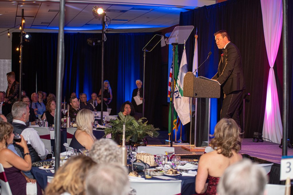 A person standing at a podium giving a speech in front of a crowd seated at round dining tables at the Mayor's Gala