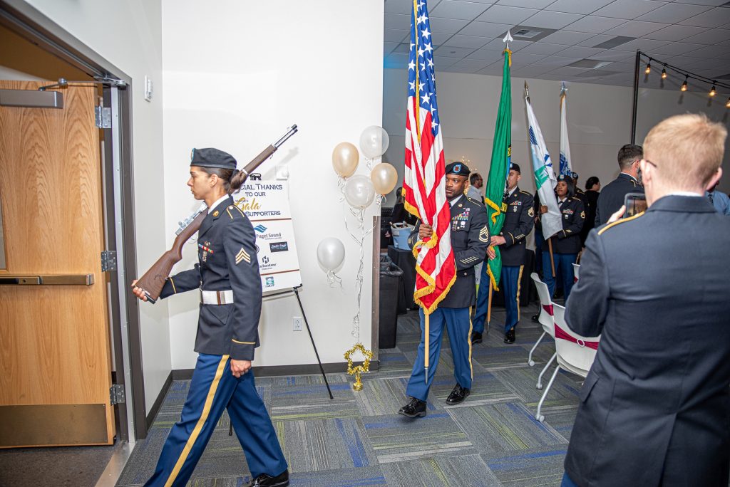 people in uniform carrying the U.S. and Washington State flag through a building, the lead person has a fake rifle over her shoulder.