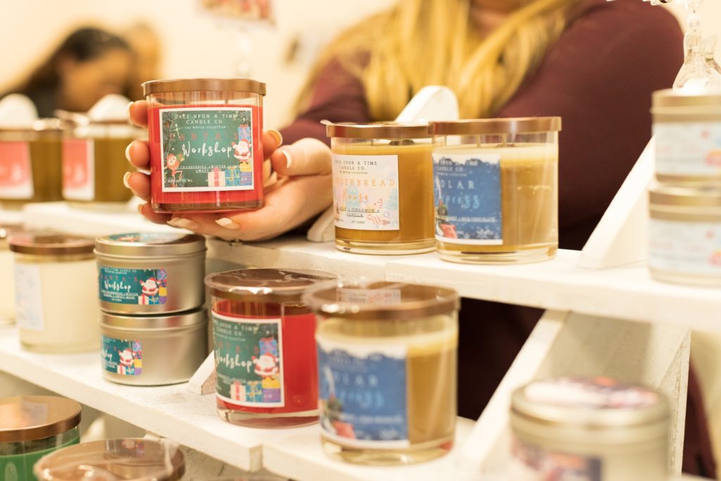 jars of candles sitting on white shelves, a woman holds one candle in her outstretched hands.