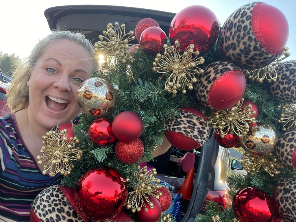 woman smiling as she holds up a large red and gold holiday wreath with cheetah print ornaments