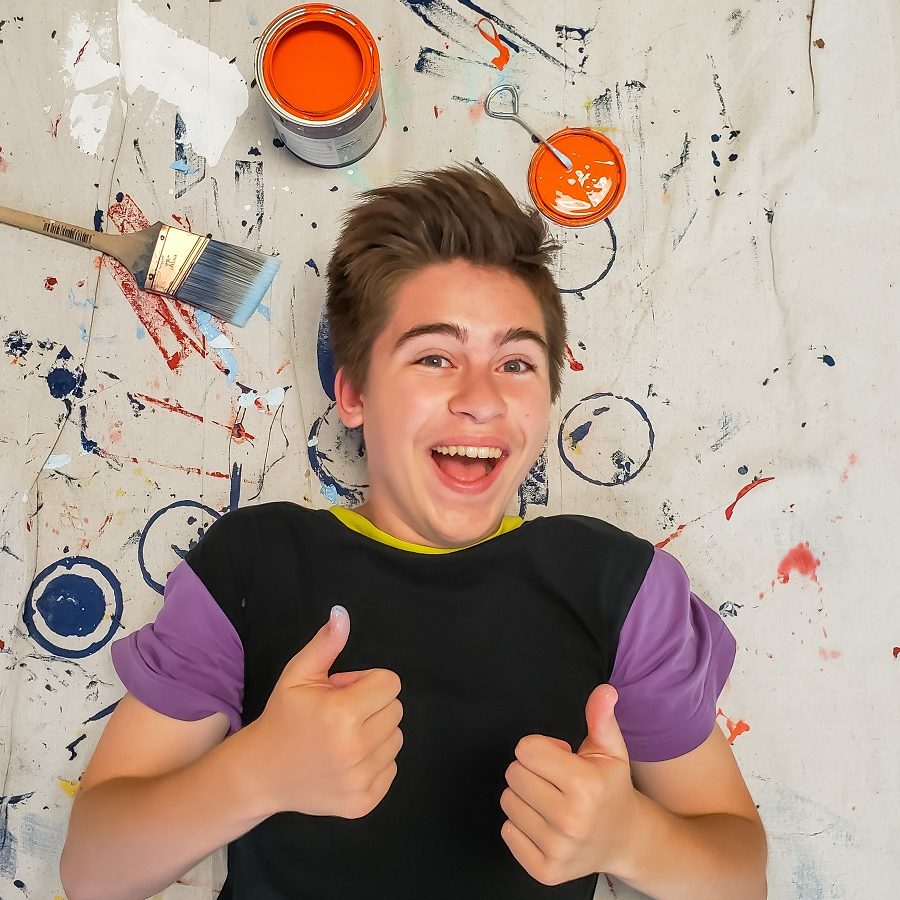 Maverick Collins lying on white fabric covered in paint swirls and paint cans. He is smiling and giving two thumbs up.